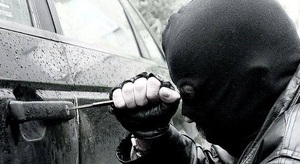 Mężczyzna w czarnej kominiarce, który śrubokręt wkręca w zamek umieszczony w drzwiach samochodu osobowego