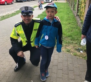 Fotografia kolorowa - Policjant zabrzańskiej drogówki na chodniku przed Przedszkolem nr 12 wręczający medal przedszkolakom biorącym udział w akcji "Jestem bezpieczny - jeżdżę w foteliku"