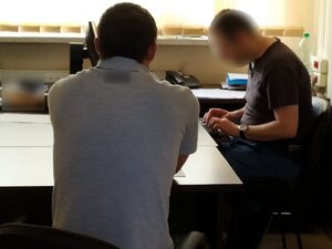 Zdjęcie kolorowe: policyjny pokój przesłuchań, podejrzany mężczyzna o pobicie dwóch przechodniów siedzący na krześle składający wyjaśnienia w obecności nieumundurowanego policjanta służby kryminalnej