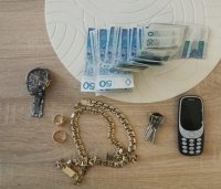 Zdjęcie kolorowe: na stole leży telefon komórkowy, zegarek, banknoty, złota biżuteria i klucze