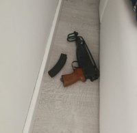 Zdjęcie kolorowe: na podłodze koło łóżka leży pistolet maszynowy skorpion