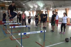 Zdjęcie kolorowe: sala gimnastyczna na które rozłożone są płotki atletyczne, policjant w stroju sportowym demonstruje jak prawidłowo pokonać tor