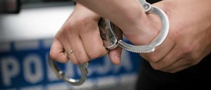 Zdjęcie kolorowe: ręce z założonymi kajdankami na tle napisu Policja