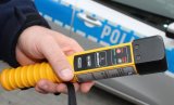 zdjęcie kolorowe: na tle policyjnego radiowozu dłoń trzymająca urządzenie do badania zawartości alkoholu w organizmie