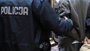 Zdjęcie kolorowe: umundurowany policjant stojacy tyłem, który trzyma zatrzymanego mężczyzne za ramię