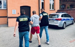 Zdjęcie kolorowe: policjanci kryminalni z IV komisariatu prowadzą zatrzymanego mężczyzne na przesłuchanie, z prawej strony stoi zaparkowany oznakowany radiowóz