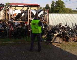 Zdjęcie kolorowe: technik kryminalistyczny w zielonej kamizelce z napisem Policja podczas oględzin ujawnionych części samochodowych