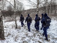 Policjanci w terenie porośniętym zaroślami, w zimowej pogodzie, w tracie poszukiwań osoby zaginionej.