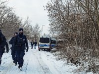 Policjanci idą drogą, w terenie  porośniętym zaroślami, w zimowej pogodzie. W tle policyjne radiowozy.