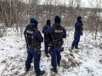 Policjanci w terenie porośniętym zaroślami, w zimowej pogodzie, w tracie poszukiwań osoby zaginionej.