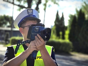 Zdjęcie kolorowe przedstawiające umundurowanego policjanta trzymającego urządzenie do pomiaru prędkości.