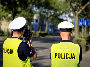 Zdjęcie kolorowe przedstawiające dwóch umundurowanych policjantów. Jeden z policjantów trzyma urządzenie do pomiaru prędkości.