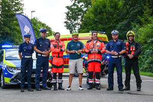 Na zdjęciu znajduje się siedem osób, w tym na środku mężczyzna w kasku oraz umundurowani policjanci i ratownicy medyczni. W tle znajdują się oznakowany radiowóz Policji oraz karetka i motocykl pogotowia ratunkowego.