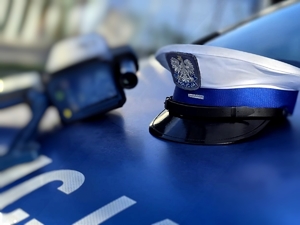 Zdjęcie kolorowe przedstawiające policyjną czapkę oraz urządzenie do ręcznego pomiaru prędkości na pokrywie silnika radiowozu.